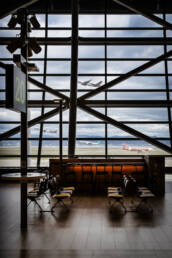 Image of airport terminal interior overlooking the runway, featuring empty seating and a bar area. large windows reveal two airplanes taking off under cloudy skies, highlighting air travel dynamics. ideal for travel enthusiasts and aviation industry insights.