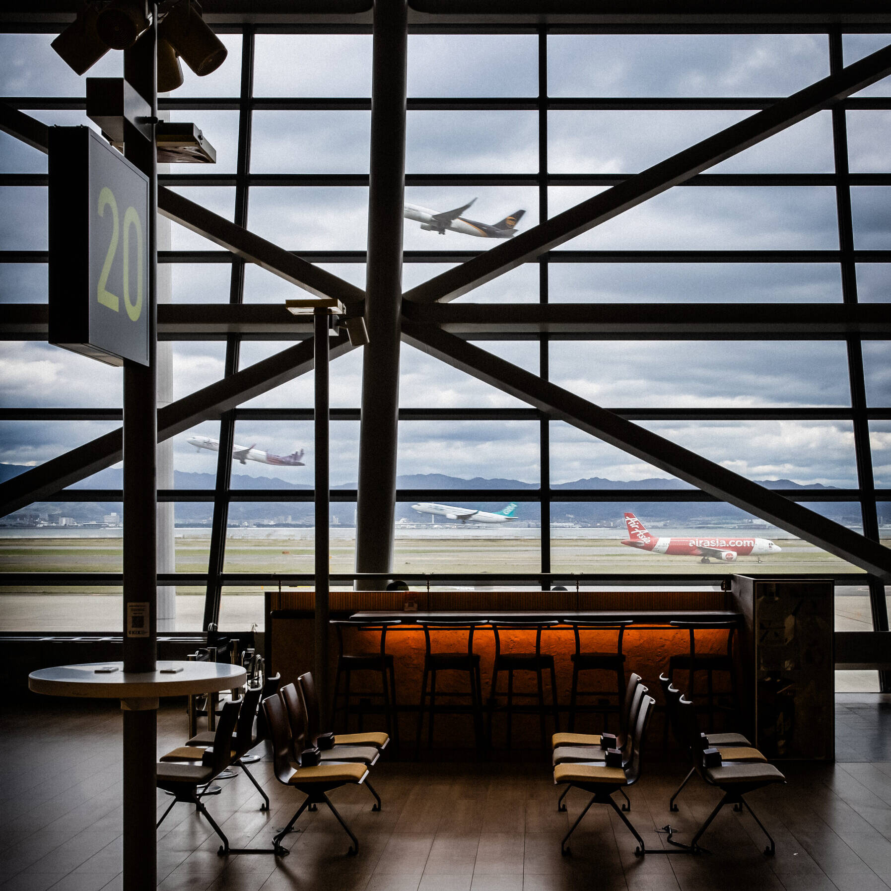 Image of airport terminal interior overlooking the runway, featuring empty seating and a bar area. large windows reveal two airplanes taking off under cloudy skies, highlighting air travel dynamics. ideal for travel enthusiasts and aviation industry insights.