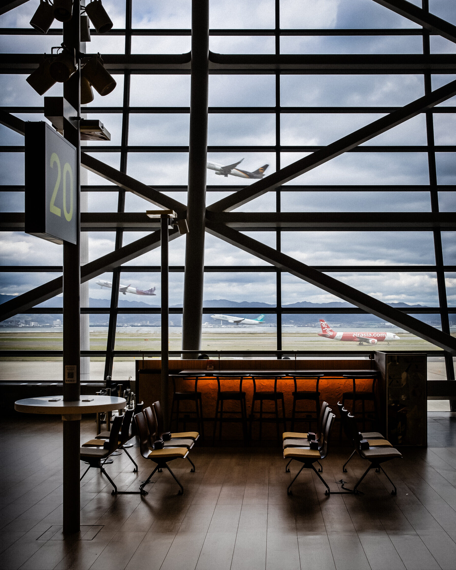 Image of airport terminal interior overlooking the runway, featuring empty seating and a bar area. large windows reveal two airplanes taking off under cloudy skies, highlighting air travel dynamics. ideal for travel enthusiasts and aviation industry insights.