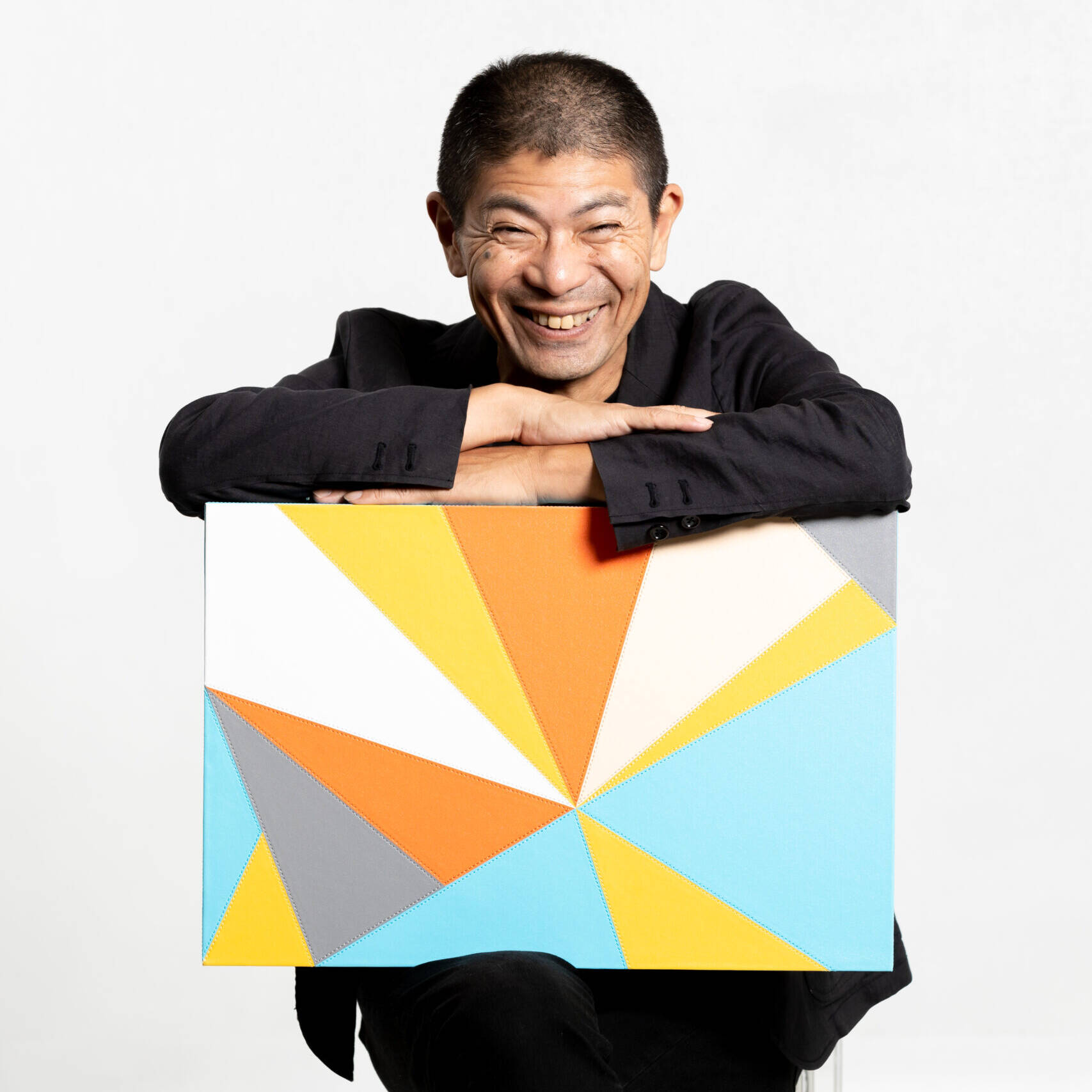 Image of smiling man in a black suit poses with a colorful geometric art piece featuring vibrant triangles in orange, blue, yellow, and gray against a neutral background. elegant and modern, this image captures the essence of contemporary art and creative expression.