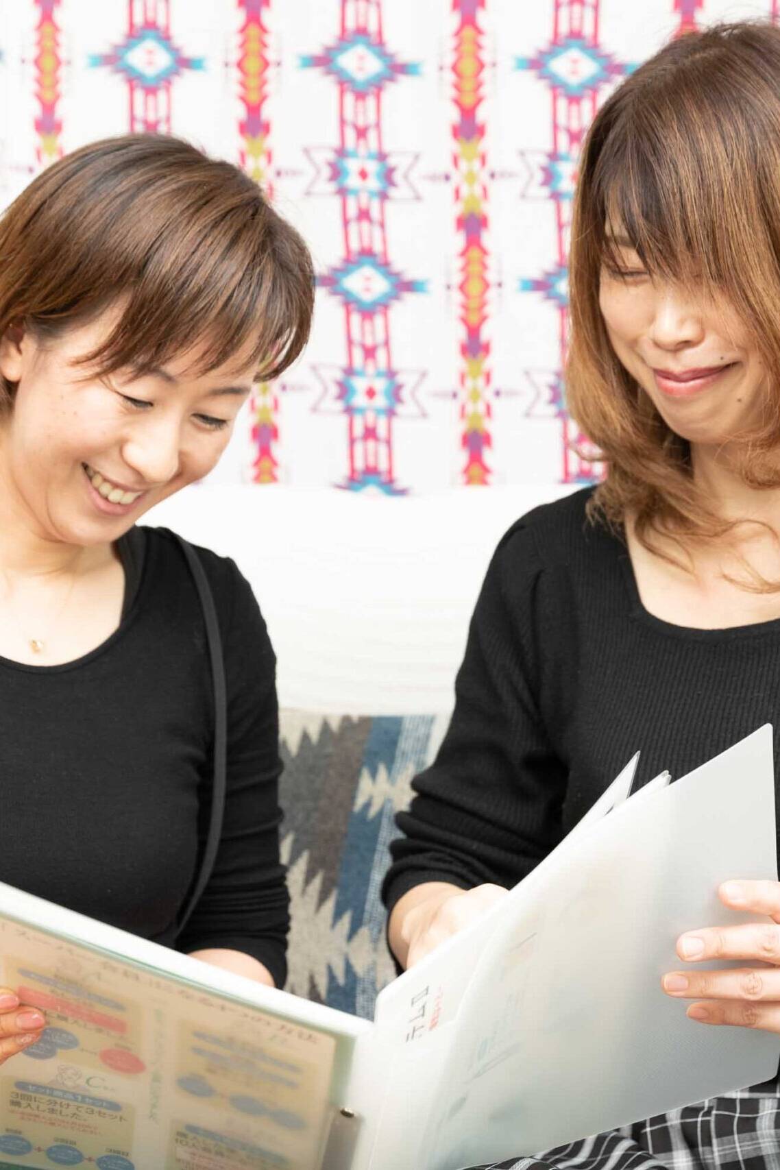 Image of two women are sitting together in a cozy living room, smiling and looking at a book or photo album. they are both wearing black tops, and there is a colorful patterned tapestry hanging on the wall behind them, adding a vibrant backdrop to the warm,