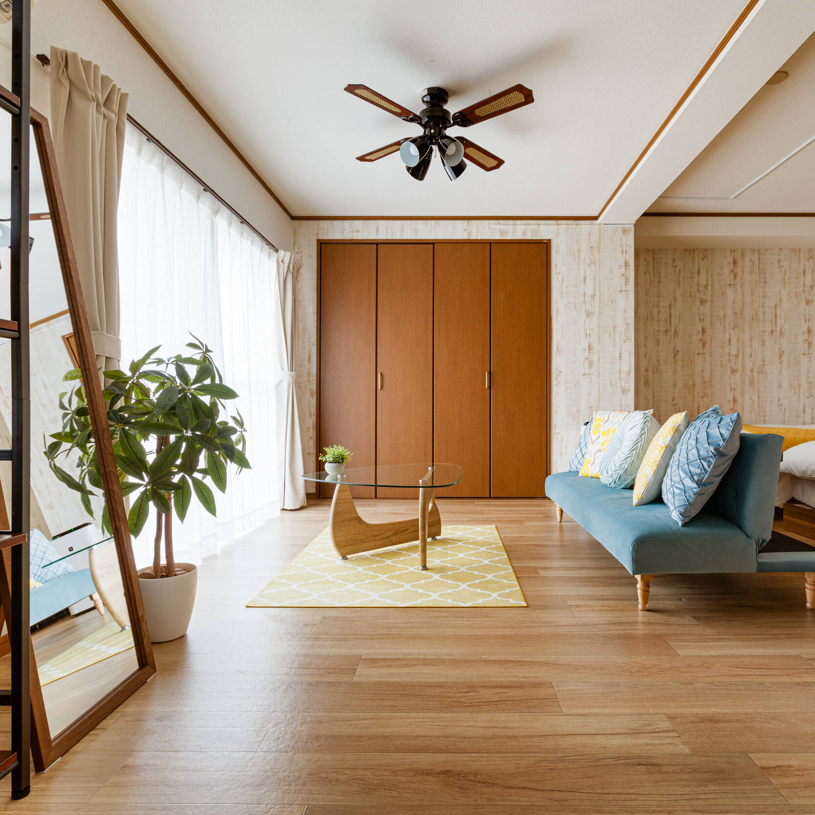 Image of this image showcases a modern, minimalist living room in a cozy apartment. the space features hardwood flooring, a stylish light blue sofa with colorful cushions, and a wooden coffee table on a yellow geometric rug. large windows with white curtains allow natural light to flood