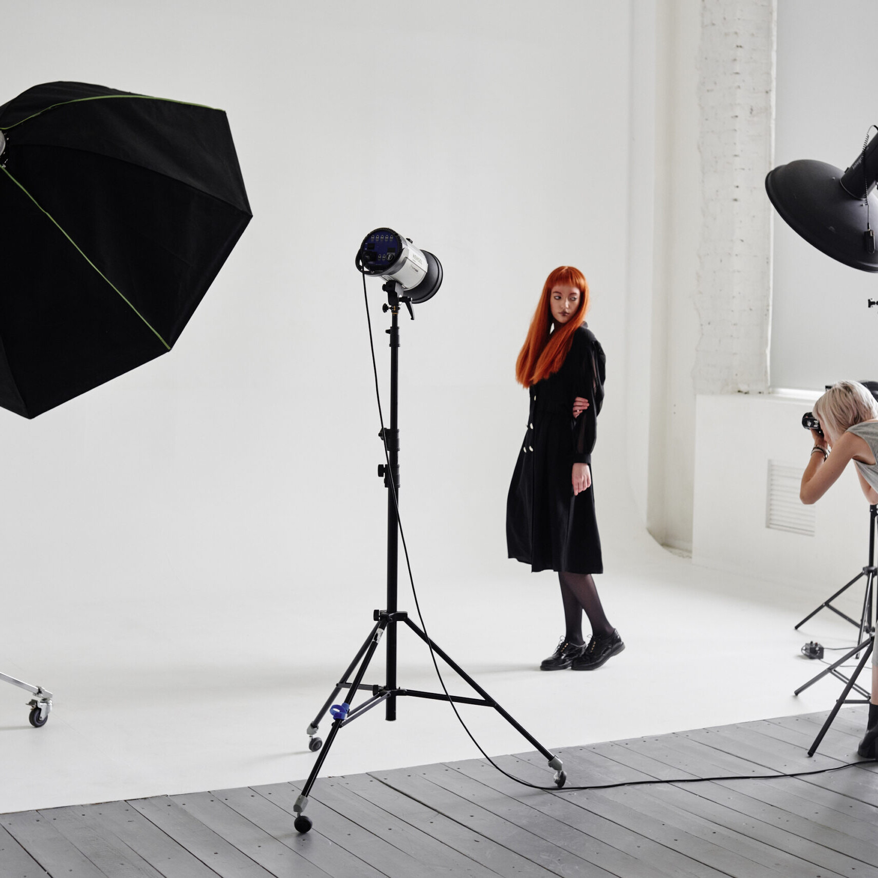 Image of fashion photography shoot in a modern studio setting with professional lighting. a photographer captures a model with long red hair and a black outfit against a white backdrop. the scene includes large softbox lighting equipment, emphasizing the creative and professional atmosphere of a fashion studio photo