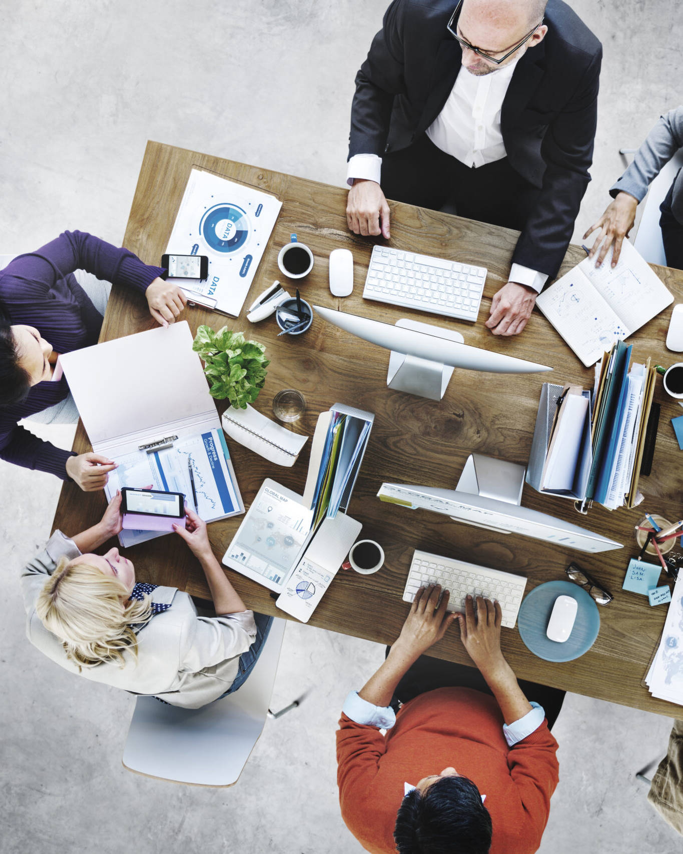 Image of overhead view of a diverse team collaborating in a modern office workspace. the image shows professionals around a wooden table engaged in discussions, using laptops, tablets, and paperwork with charts and graphs. ideal for keywords like
