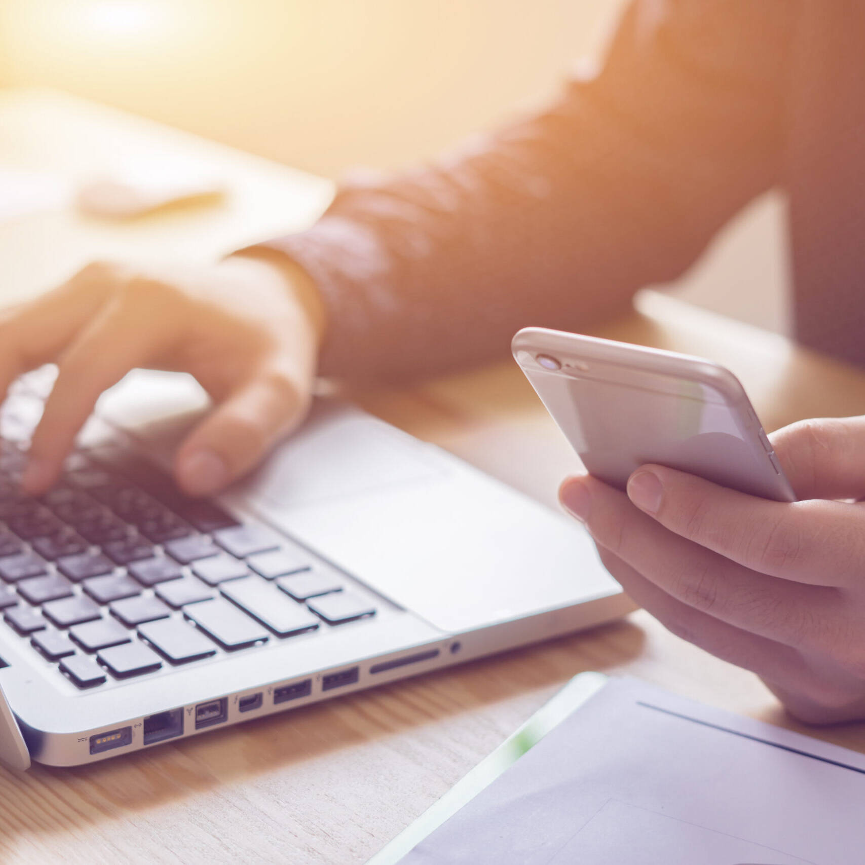 Image of a person is multitasking with technology, using a laptop and smartphone at a desk, highlighting modern digital workspace, productivity, and remote work solutions.