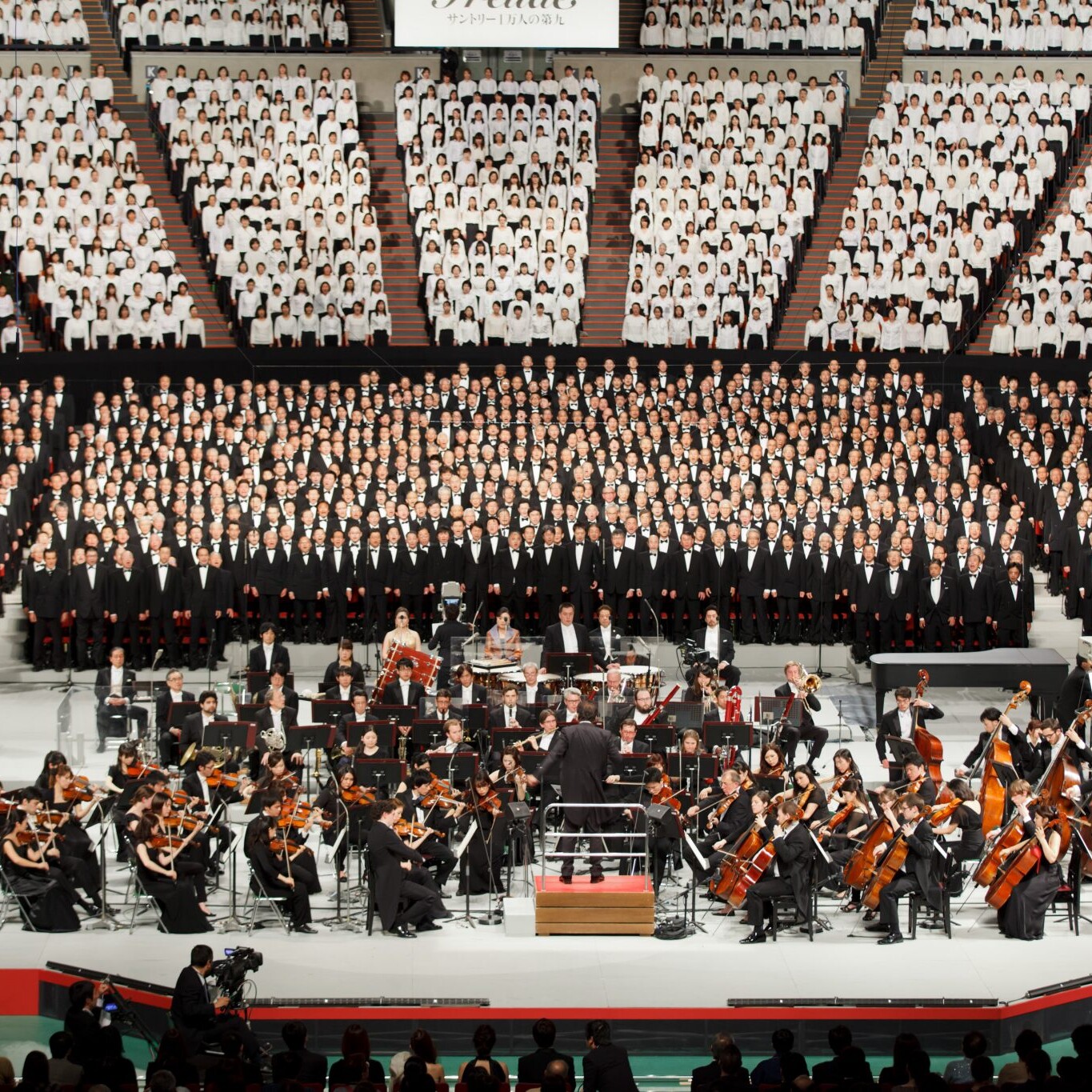 Image of this image captures a grand choral performance featuring a massive choir and full orchestra in a large concert venue. the choir, dressed in black suits and white shirts, stands behind the orchestra, which includes violinists, cellists, and other musicians. the