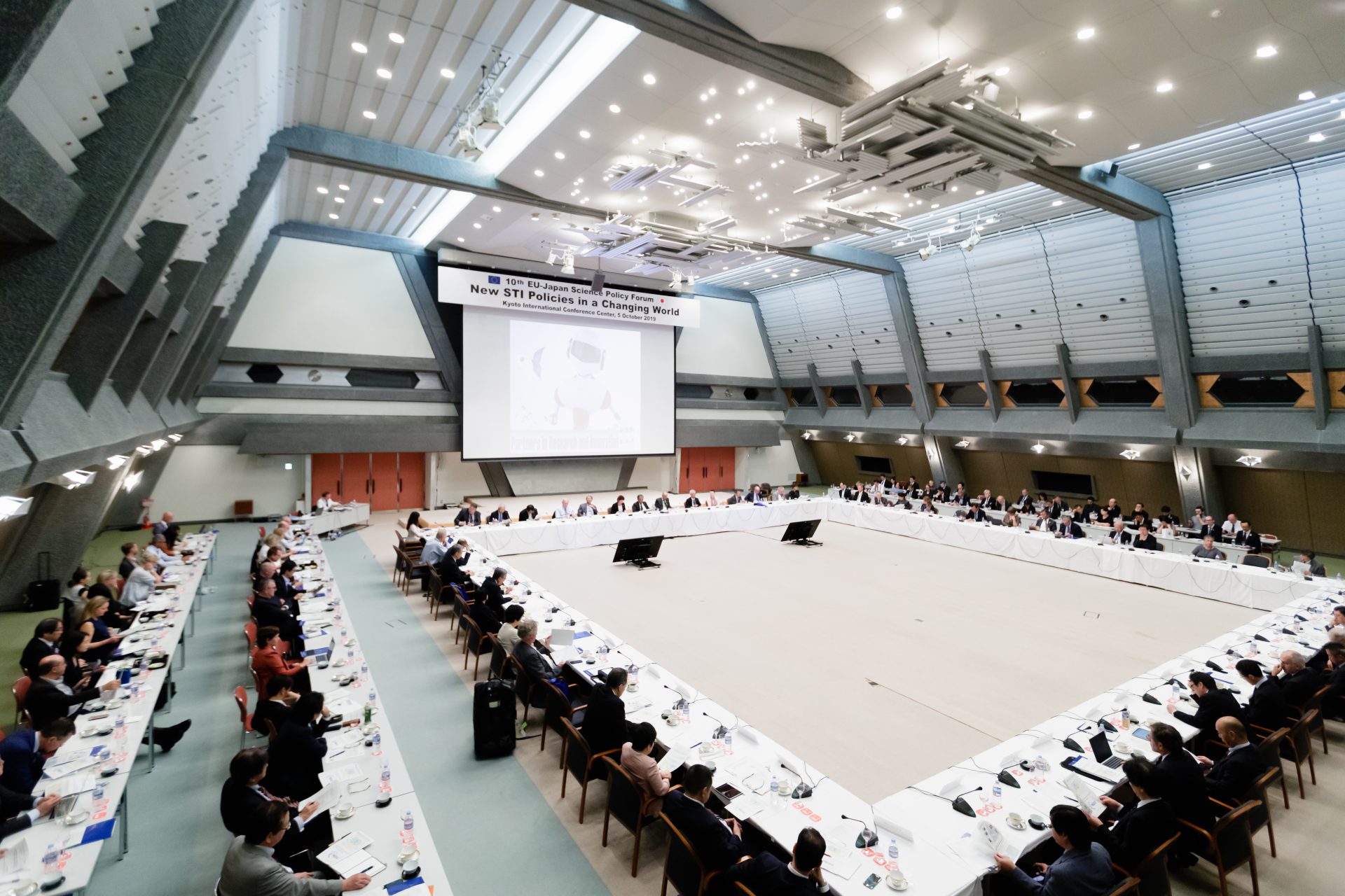 Image of the image showcases the 10th eu-japan scientific policy forum held at the kyoto international conference center. the spacious conference room features a large round table with numerous international delegates engaged in discussions. the event focuses on 