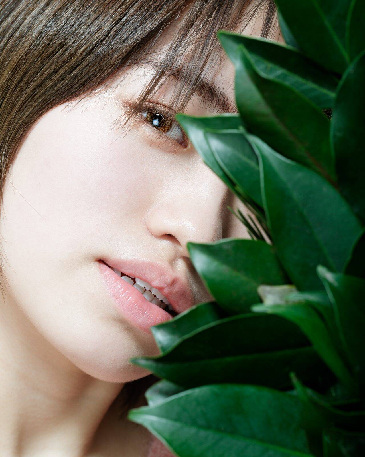 Image of portrait of a young woman with a close-up view, partially obscured by lush green leaves. the focus on her natural beauty and delicate features creates a serene and organic atmosphere, perfect for themes related to nature, skincare, and minimalist aesthetics.
