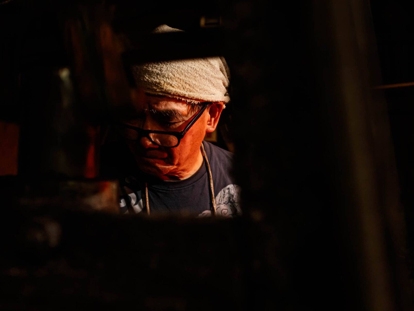 Image of the image depicts an elderly man working in dim lighting, suggesting a traditional workshop setting. he wears glasses and a headscarf, concentrating on a task. this photo captures the essence of craftsmanship and dedication in an artisanal environment, highlighting themes of skilled