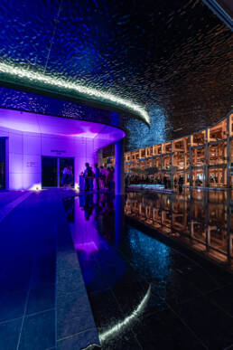 Image of a stunning architectural scene featuring a modern entrance lit with vibrant purple and blue lights, reflecting elegantly on a sleek water feature. the meticulously designed space showcases a mix of contemporary and futuristic elements, creating an inviting ambiance. this artistic indoor setup is ideal for night