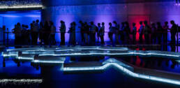 Image of silhouetted against a vibrant blue and purple backdrop, a group of people wait in line inside a modern exhibition. the scene is illuminated by reflective led floor lighting, adding a futuristic ambiance. ideal for keywords: indoor lighting, modern exhibit, vibrant colors