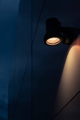 Image of modern outdoor wall light fixture illuminating a sleek, dark building facade at dusk. the warm glow contrasts with the deep blue evening sky, highlighting the architectural design. perfect for showcasing urban lighting solutions and contemporary building aesthetics. keywords: outdoor lighting, modern architecture,