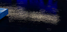 Image of rainwater splashes create ripples on a wet, reflective surface, illuminated by dramatic blue and white lighting. the dark water captures the essence of a rainy urban evening, highlighting the interplay of light and texture for a captivating visual effect. keywords: rain