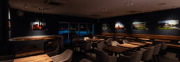 Image of modern, cozy cafe interior with elegant seating and wooden tables, featuring dim ambient lighting. the walls are adorned with vibrant landscape photography, creating an art gallery ambiance. large windows provide views of the outdoor terrace, enhancing the open, inviting atmosphere. ideal for