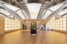 Image of modern airport interior featuring sleek architectural design, showcasing prominent ceiling beams and ambient lighting. central signage directs passengers to departures and escalators. the spacious wooden-floor terminal emphasizes efficient navigation and contemporary travel amenities.