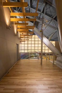 Image of a modern architectural interior featuring a wooden walkway with ceiling beams, leading to an illuminated wall and an escalator. the design incorporates industrial elements, warm lighting, and a sleek, contemporary aesthetic, ideal for a transit or commercial space. keywords: modern architecture