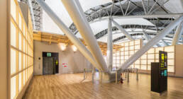 Image of this image captures the modern interior of an airport terminal, showcasing its sleek architectural design with large metal beams and a wooden floor. illuminated signage directs travelers towards departure areas, emphasizing accessibility and convenience in a bustling transportation hub. the bright lighting and open structure