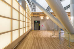 Image of this image showcases a modern, well-lit indoor area with polished wooden flooring, large illuminated wall panels, and structural beams. the space includes an accessible entrance with a wheelchair-friendly sign, suitable for public venues such as airports or shopping malls. the contemporary design