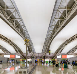 Image of the image depicts the interior of a modern airport terminal, showcasing its sleek architectural design. the spacious hall is lined with large, curved ceilings supported by steel beams, creating an open and airy atmosphere. passengers with luggage are visible, engaging in typical airport