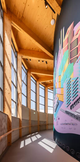 Image of this image showcases a modern architectural interior featuring a high-ceilinged space with large windows, allowing abundant natural light to flood the area. the wood beams and contemporary design elements are accented by a vibrant, colorful mural depicting an urban scene with buildings and trains