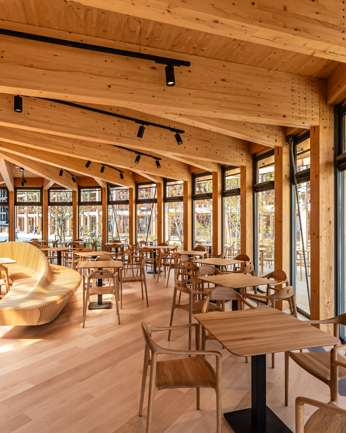 Image of this image showcases the interior of a modern wooden cafe, featuring an open and airy design with large windows allowing ample natural light. the space is furnished with sleek wooden tables and chairs, complemented by a curved wooden bench. the angled wooden beams of the ceiling