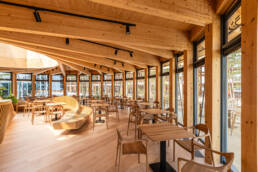 Image of this image showcases the interior of a modern wooden cafe, featuring an open and airy design with large windows allowing ample natural light. the space is furnished with sleek wooden tables and chairs, complemented by a curved wooden bench. the angled wooden beams of the ceiling