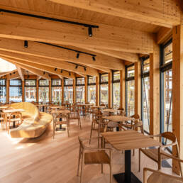 Image of this image showcases the interior of a modern wooden cafe, featuring an open and airy design with large windows allowing ample natural light. the space is furnished with sleek wooden tables and chairs, complemented by a curved wooden bench. the angled wooden beams of the ceiling