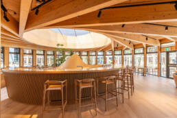 Image of this image showcases a modern cafe interior with a wooden circular bar at its center, featuring a unique geometric ceiling design and ample natural light streaming through large windows. the setting includes stylish wooden stools and lush greenery, creating a warm and inviting atmosphere. ideal for