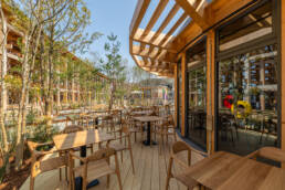 Image of this image showcases a beautifully designed outdoor cafe patio, set under a wooden pergola. the scene features modern wooden tables and chairs, surrounded by lush greenery, offering a serene dining experience. ideal for those seeking an eco-friendly and tranquil ambiance, this cafe